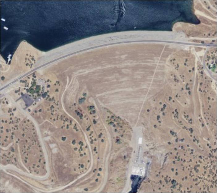A top-down USGS aerial image showing Don Pedro Dam’s broad earth-and-rock embankment between the reservoir and downstream powerhouse