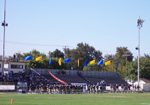 Football players and spectators beside the bleachers at Joe Debely Stadium in an older photograph.