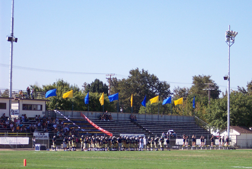 Football players and spectators beside the bleachers at Joe Debely Stadium in an older photograph.