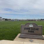 John Lazar Park sign beside a broad lawn, with blue playgrounds and surrounding homes in Turlock.