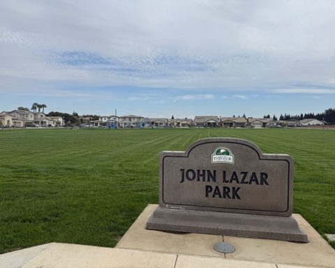 John Lazar Park sign beside a broad lawn, with blue playgrounds and surrounding homes in Turlock.